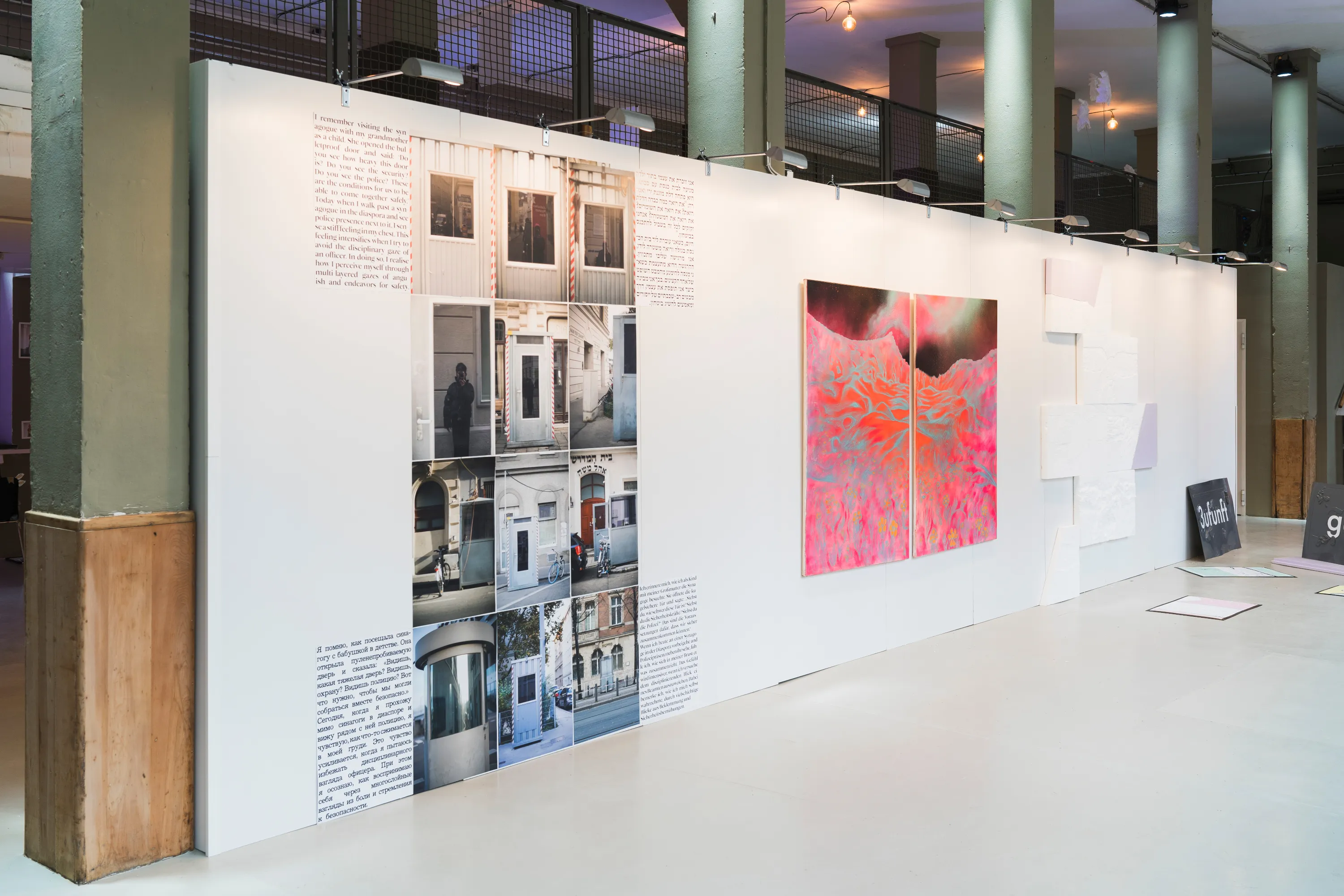 Das Bild zeigt die Ausstellungsansicht des ersten Rundgangs der Jüdischen Kunstschule. An einer weißen Wand hängen verschiedene Kunstwerke: eine wandfüllende Collage bestehend aus Fotografien und Text, zwei großformatige, abstrakte Gemälde in Rottönen sowie kleine Reliefs aus Styropor, die mit auf dem Boden liegenden Objekten eine weitere konzeptuelle Arbeit bilden.