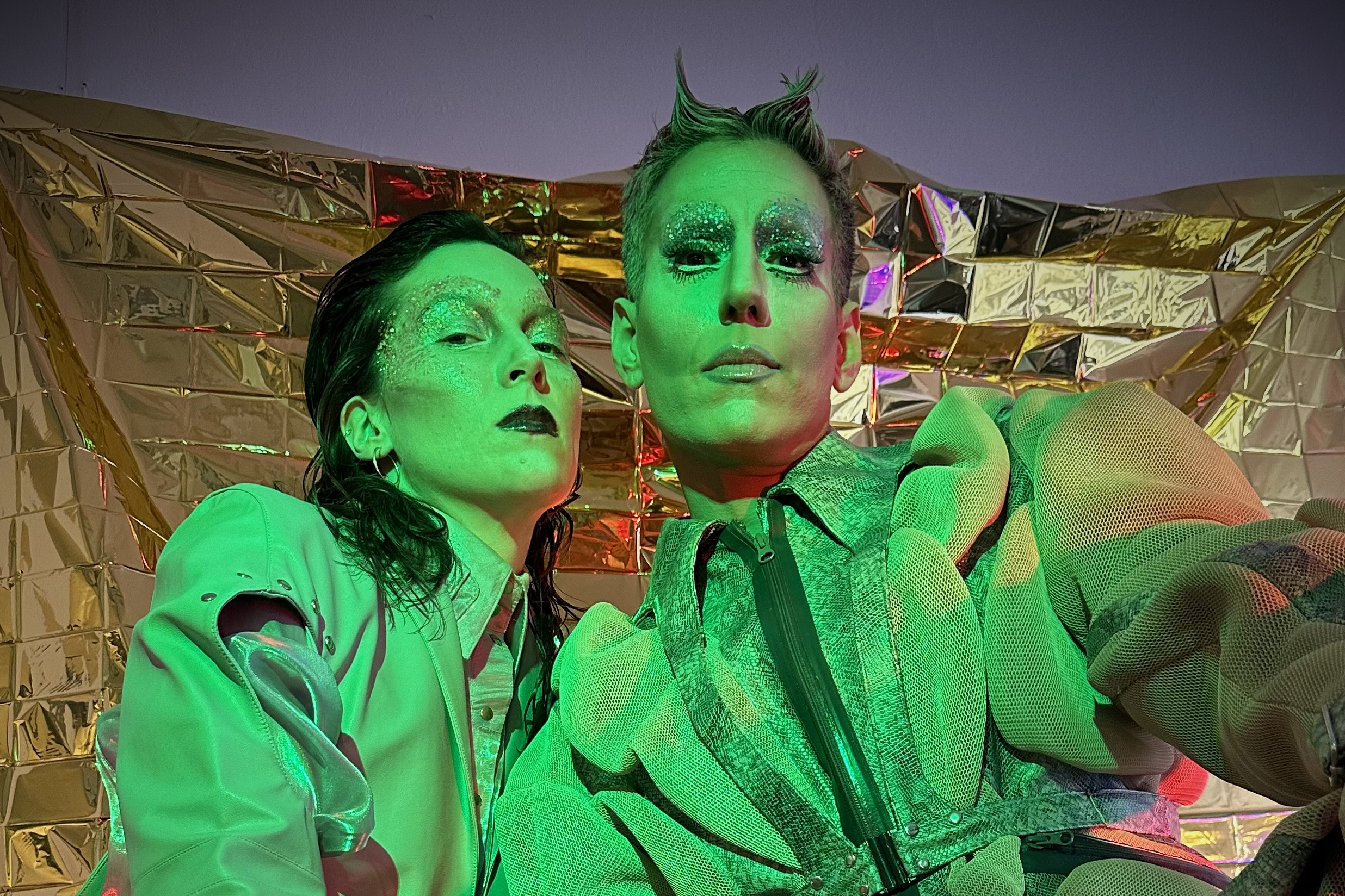 The picture shows two performers from the House of Diaspora performance series. The two pose in eye-catching, bright green outfits and wear extravagant make-up with glitter. The clothing looks avant-garde and the scene is strongly accentuated by green and red lighting.