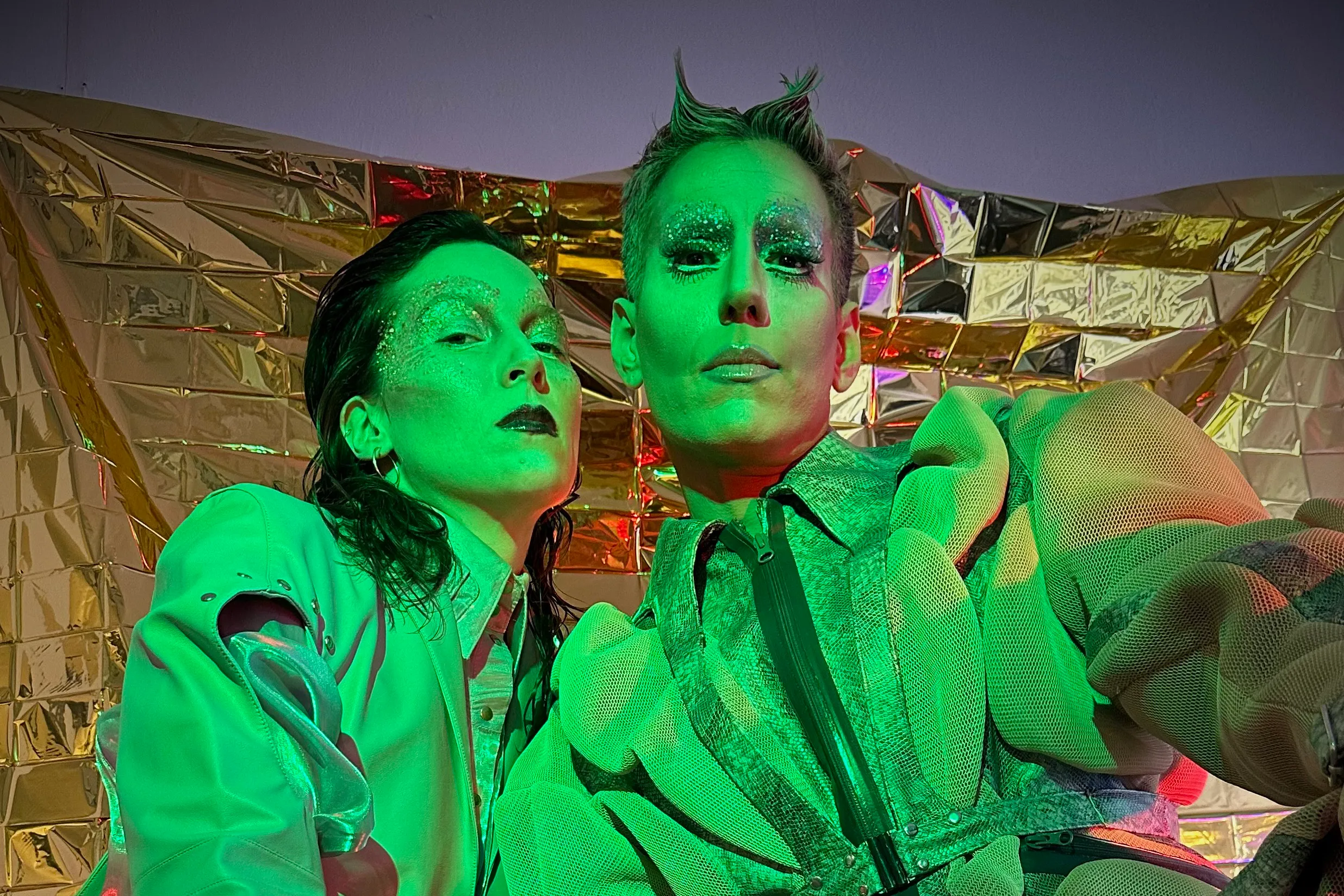 Das Bild zeigt zwei Performer:innen der Performance-Serie House of Diaspora. Die Beiden posieren in auffälligen, hellgrünen Outfits und tragen extravagantes Make-up mit Glitzer. Die Kleidung wirkt avantgardistisch und die Szene ist stark durch grüne und rote Beleuchtung akzentuiert.