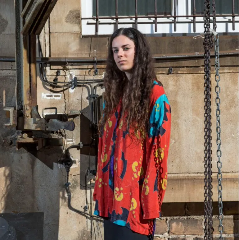 Foto von weiblich gelesener Person mit braunen langen Locken, die in die Kamera blickt. Daneben Überschrift: »Berlin ist zu meiner Realität geworden« Die Filmemacherin Shoshana Simons über ihre Arbeit, das Schtetl und die Jüdische Kunstschule