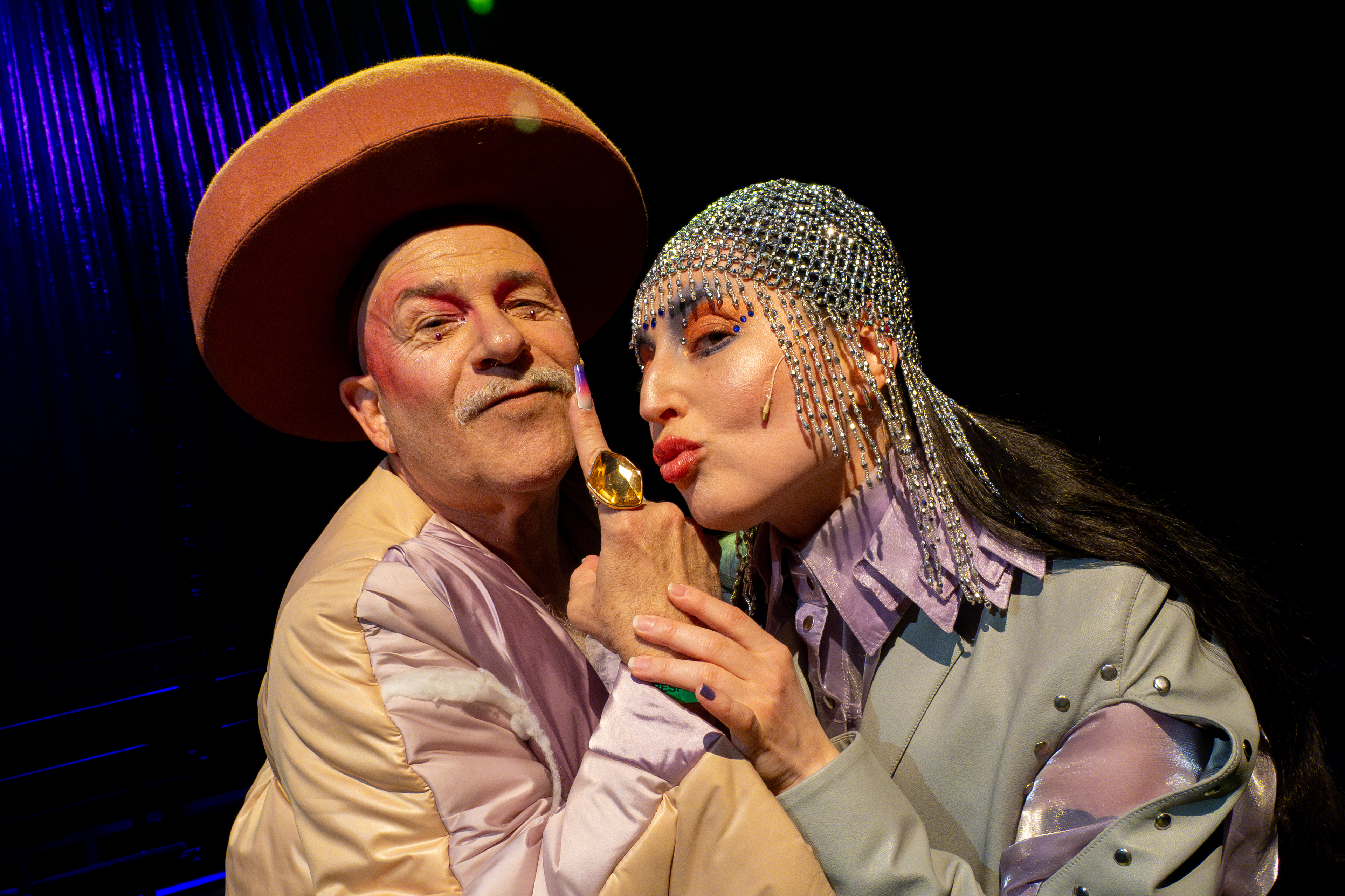 The picture was taken during Purimshpil 2025 at the Münchner Kammerspiele and presents the performance series House of Diaspora. You can see two people in eye-catching costumes, one with a large hat and the other with a glittering headdress. The characters are King Achashverosh and King Esther.