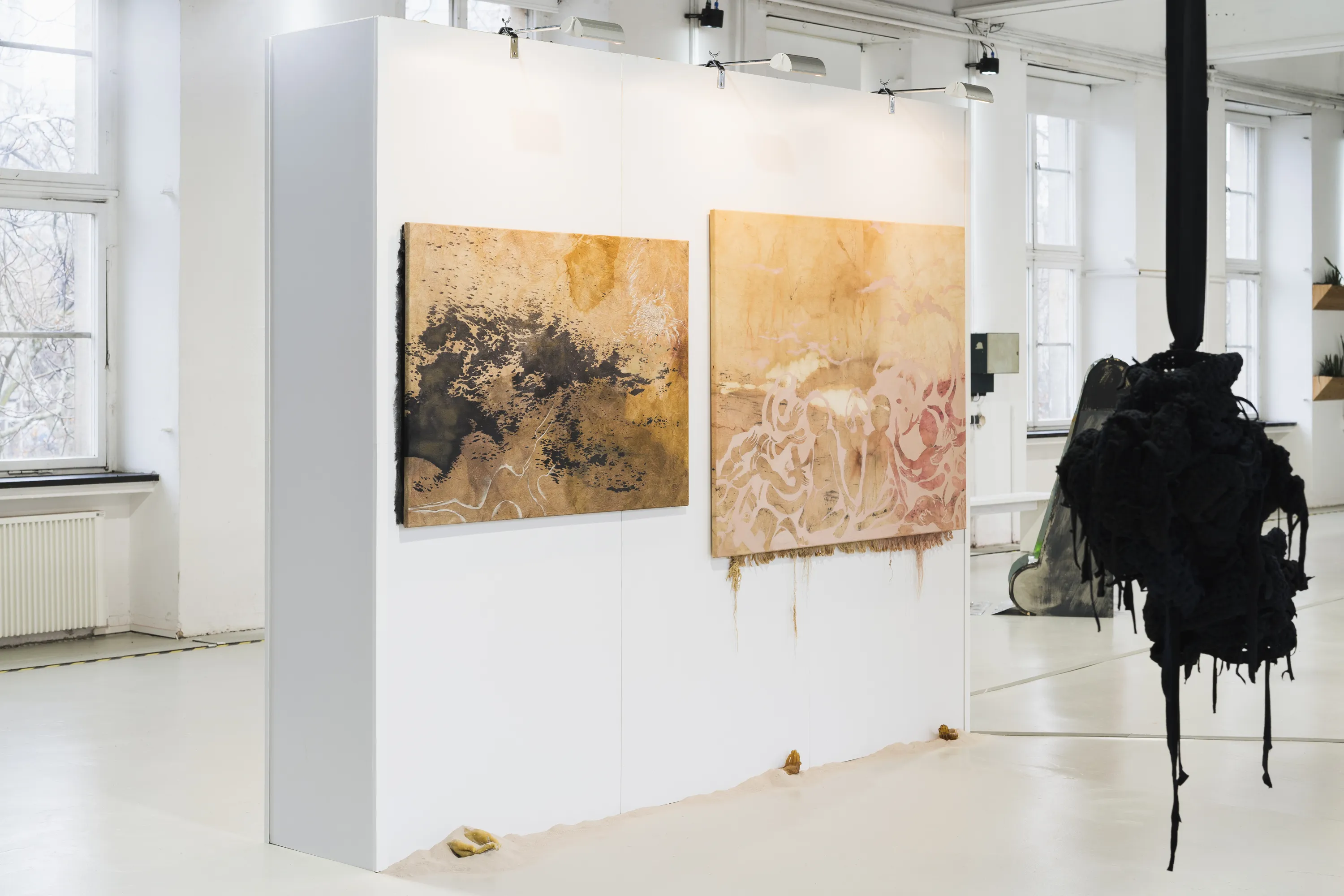 Jüdische Kunstschule Das Bild zeigt die Ausstellungsansicht des ersten Rundgangs der Jüdischen Kunstschule. Zwei abstrakte Gemälde in Erdtönen hängen an einer weißen Stellwand in hellen Ausstellungsräumen mit großen Fenstern. Im Vordergrund hängt eine organisch wirkende Skulptur von der Decke.