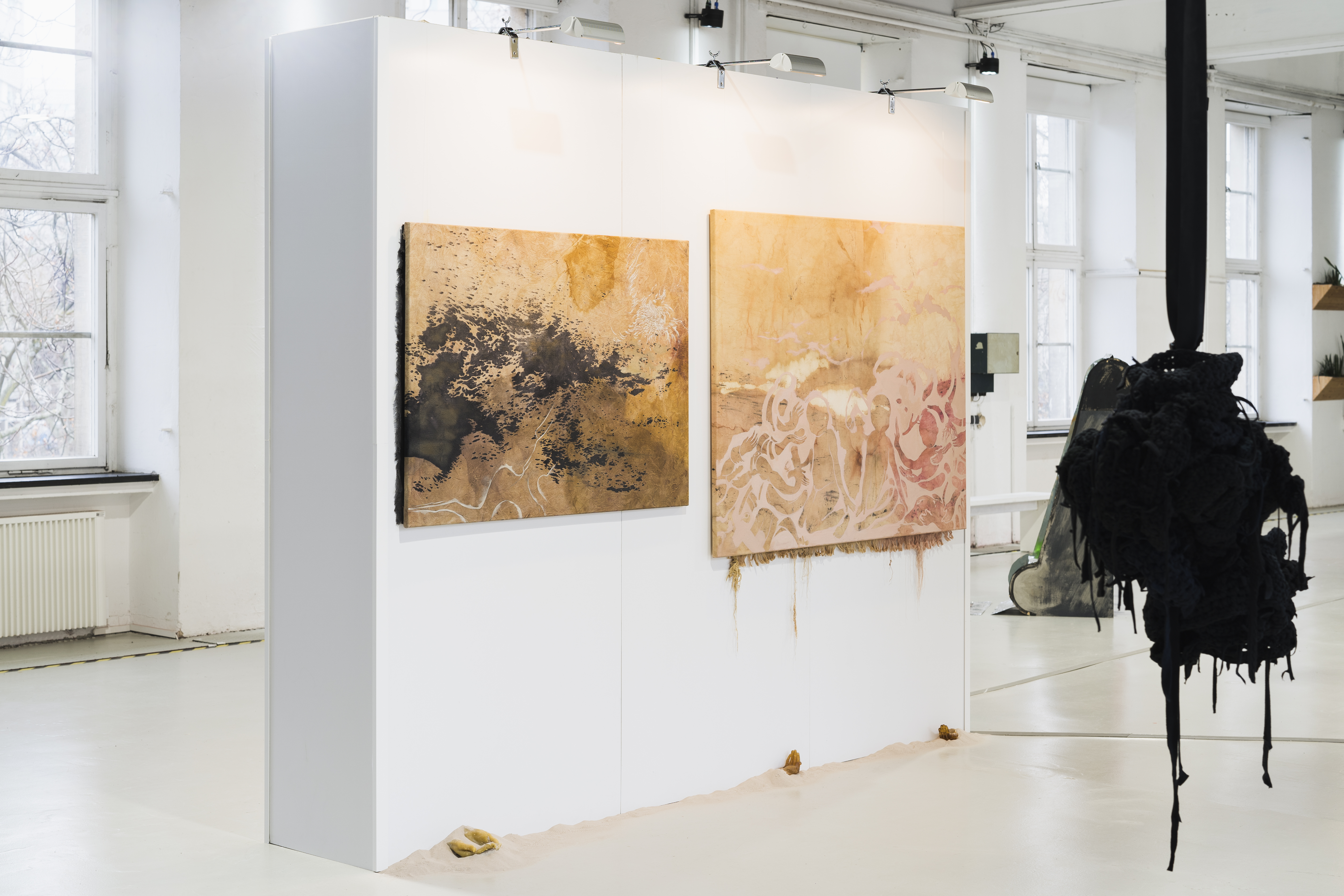 The picture shows the exhibition view of the first tour of the Jewish School of Art. Two abstract paintings in earth tones hang on a white wall in bright exhibition rooms with large windows. In the foreground, an organic-looking sculpture hangs from the ceiling.