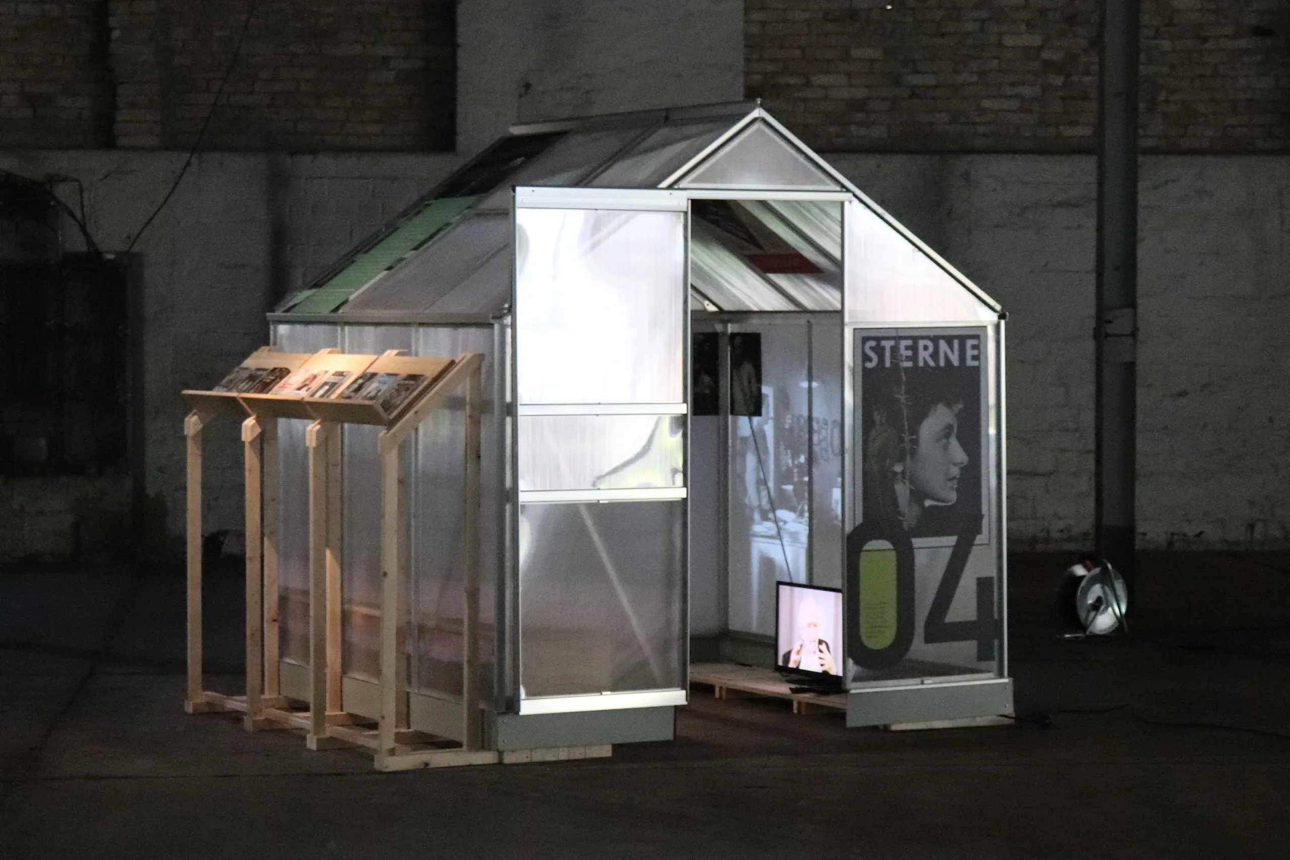 Zu sehen ist eines der kleinen Gewächshäusschen der Ausstellung im Messingwerk zu 100 Jahren jüdischer Migrationsgeschichte in Brandenburg. Das Haus, gebaut aus durchsichtigen Paneelen, steht in einer dunklen Ecke einer großen Industriehalle. Die Tür ist geöffnet. Im Haus läuft eine Videoprojektion. Außen am Haus ist ein Poster zu sehen.