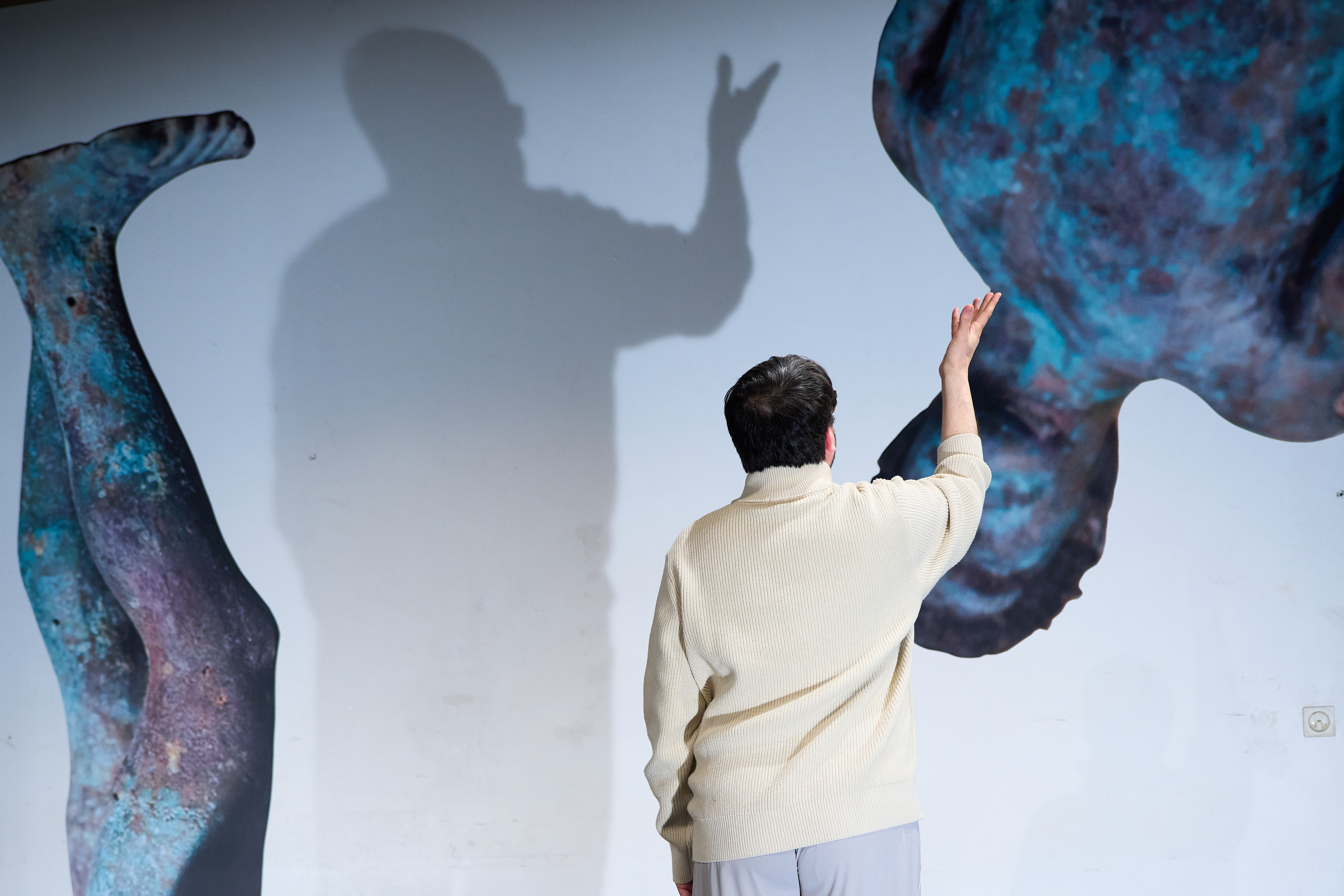 The picture was taken during the premiere of the play Dualidarity. It shows one of the two actors standing in front of a wall on which images of two large, fragmented Apollo sculptures can be seen. The actor casts a shadow on the wall, showing a hand with outstretched fingers.