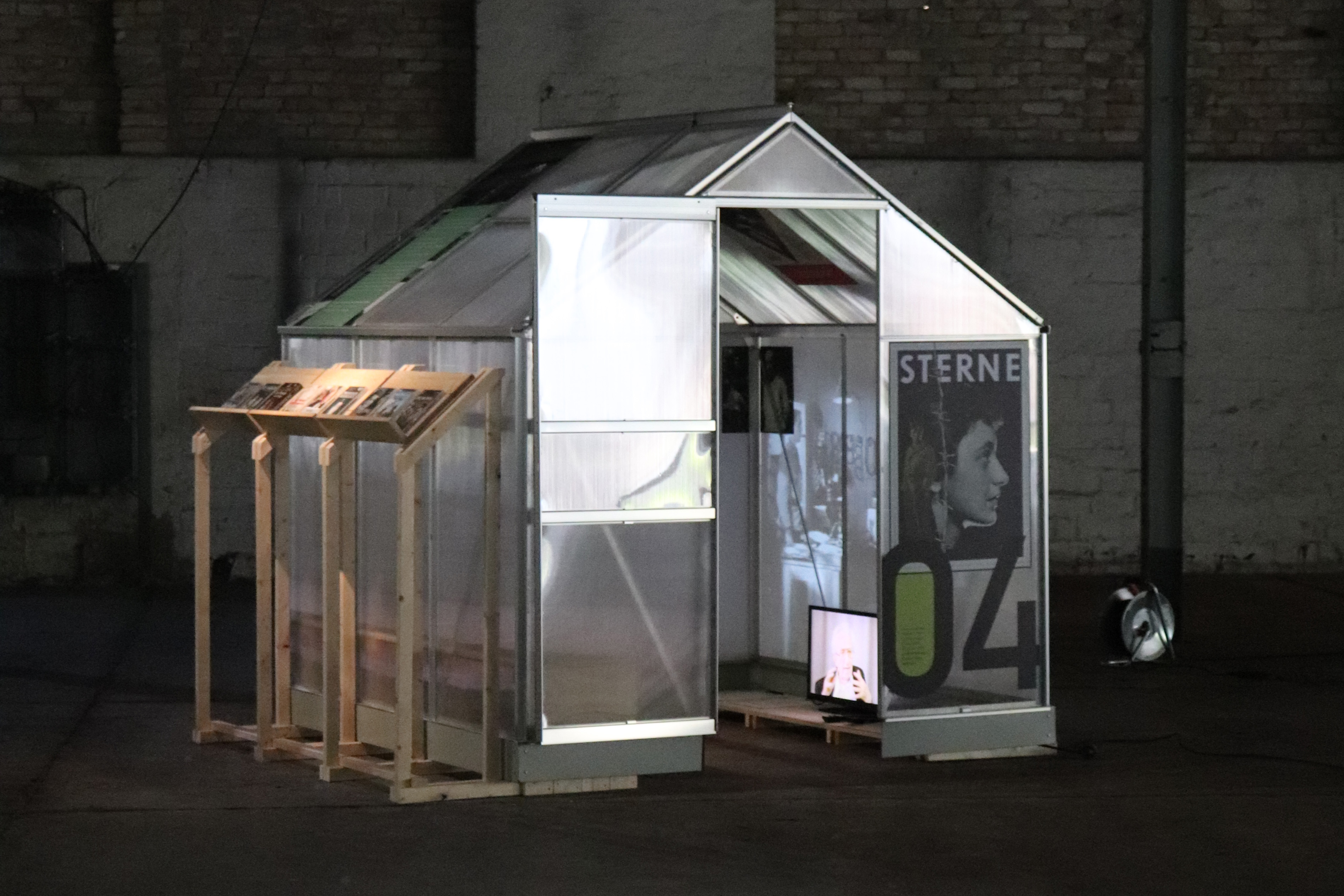 Zu sehen ist eines der kleinen Gewächshäusschen der Ausstellung im Messingwerk zu 100 Jahren jüdischer Migrationsgeschichte in Brandenburg. Das Haus, gebaut aus durchsichtigen Paneelen, steht in einer dunklen Ecke einer großen Industriehalle. Die Tür ist geöffnet. Im Haus läuft eine Videoprojektion. Außen am Haus ist ein Poster zu sehen.