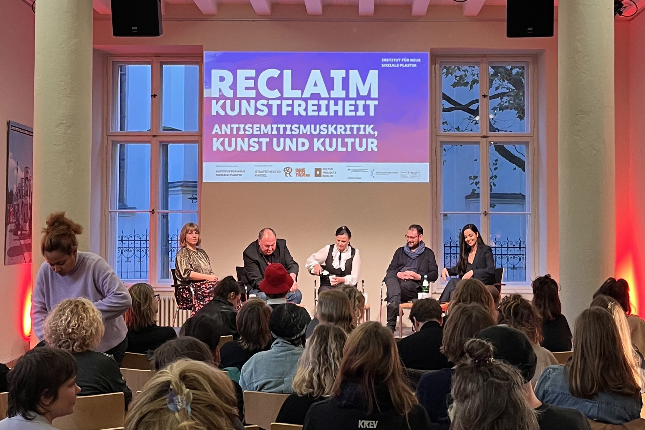 Podiumsdiskussion zur Abschlussveranstaltung vom Projekt »Reclaim Kunstfreiheit: Antisemitismuskritik, Kunst und Kultur«. Zu sehen sind fünf Personen vor Publikum, mit einer großen Projektion im Hintergrund.