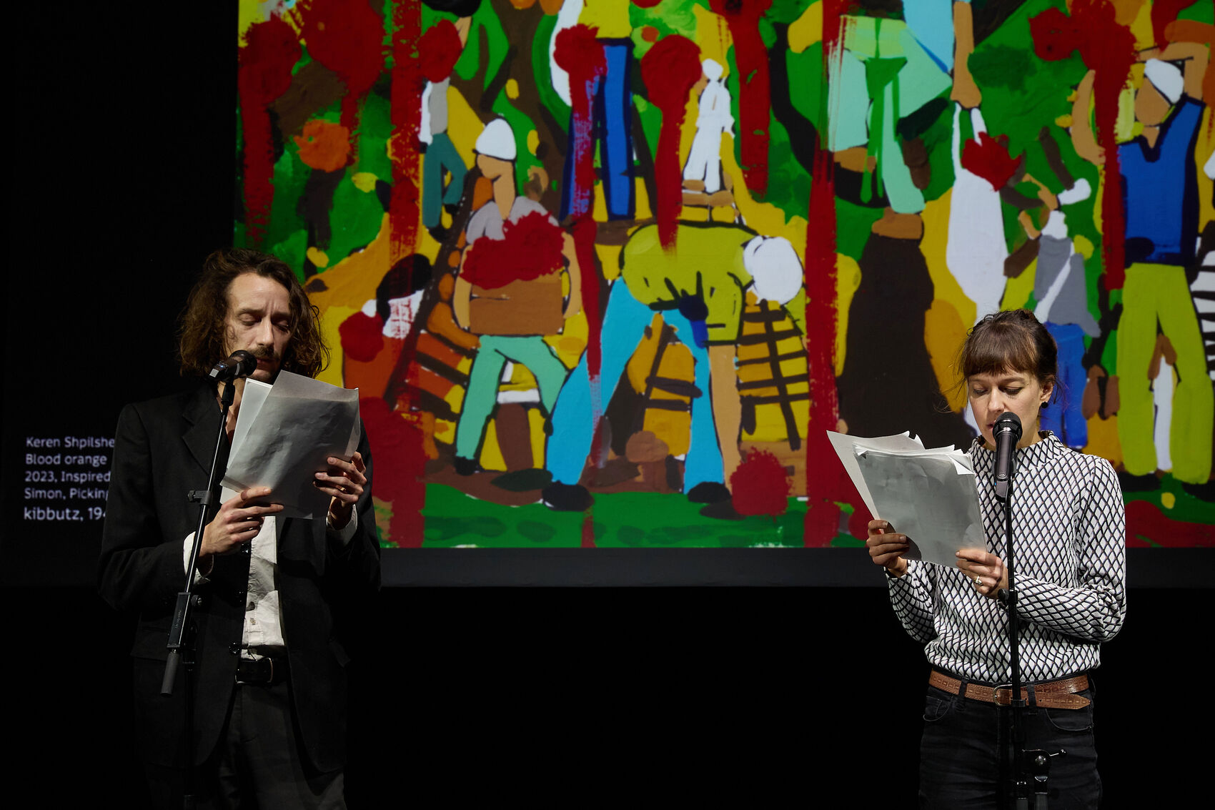 The picture shows two people standing in front of a large canvas and speaking into microphones. A colorful, expressive painting can be seen on the canvas.