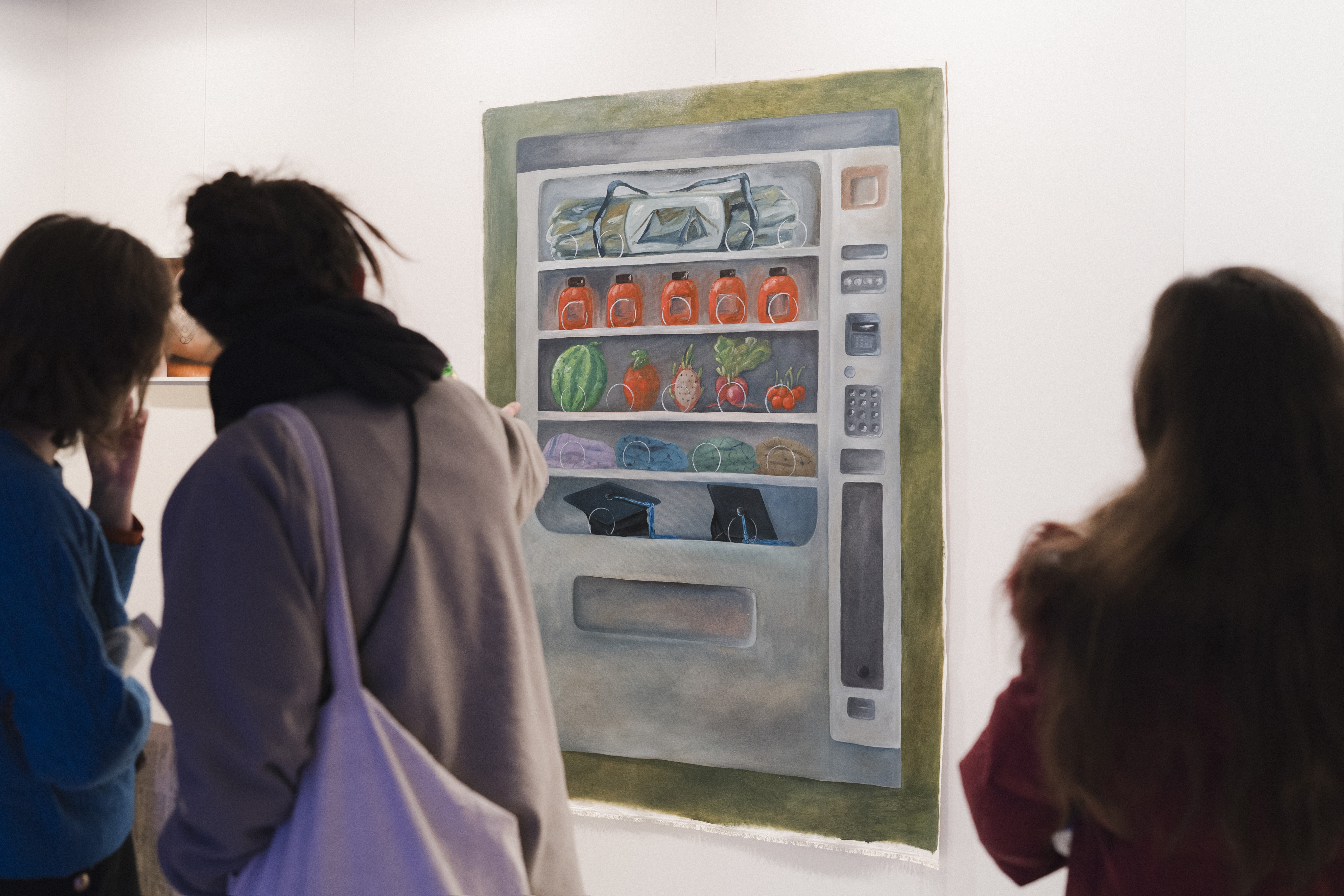Das Bild zeigt die Ausstellungsansicht des ersten Rundgangs der Jüdischen Kunstschule. Drei Personen betrachten ein Gemälde eines Verkaufsautomaten mit Wasserflaschen, Marmeladengläsern, Obst und Gemüse in einer Galerie.